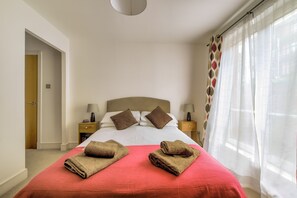 A hotel room with a bed, two bedside tables with lamps, a window with sheer curtains, and a door leading to another room.