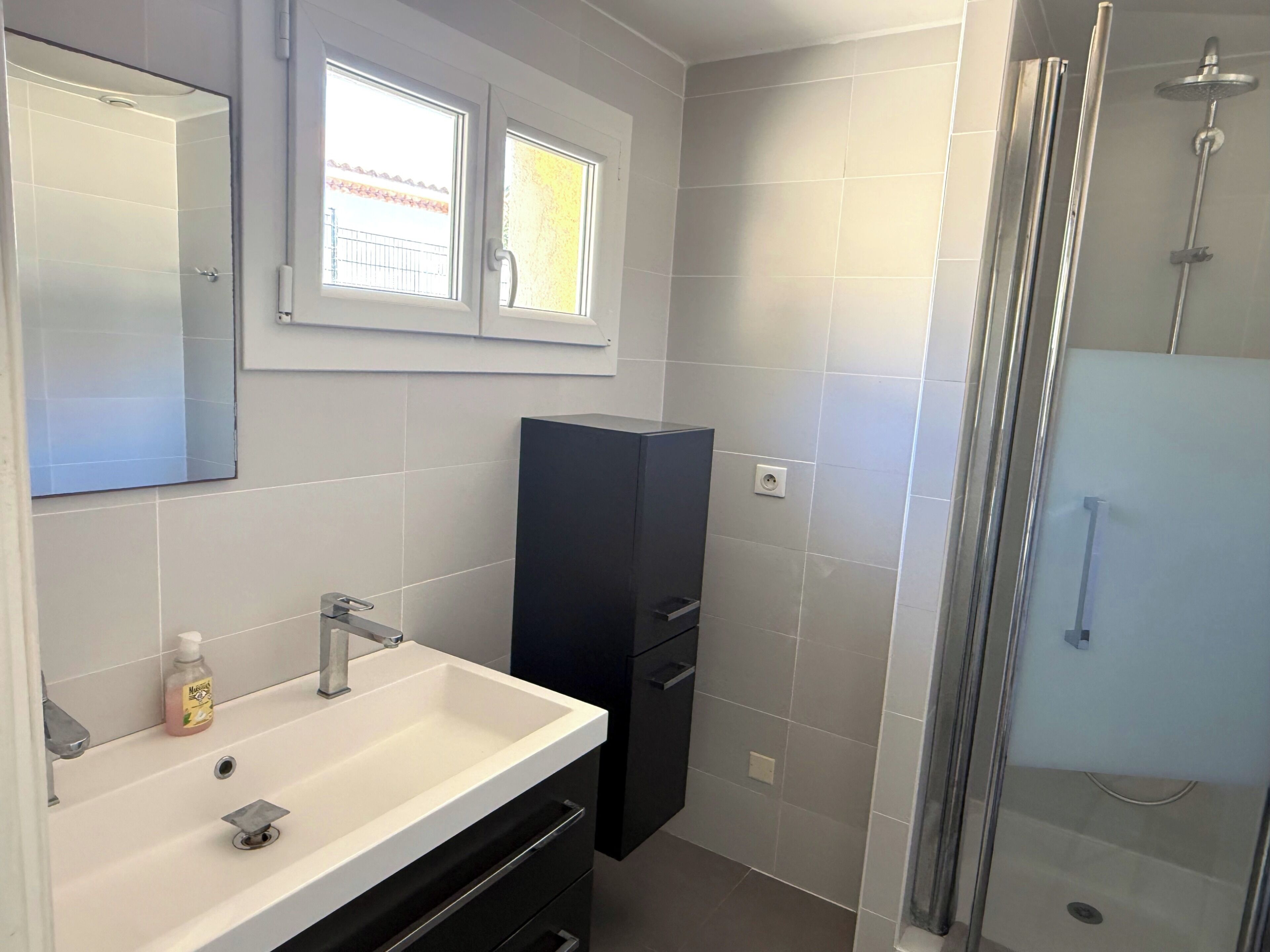 Un baño moderno con un lavamanos blanco, un espejo, un mueble oscuro y una ducha con mampara de vidrio.
