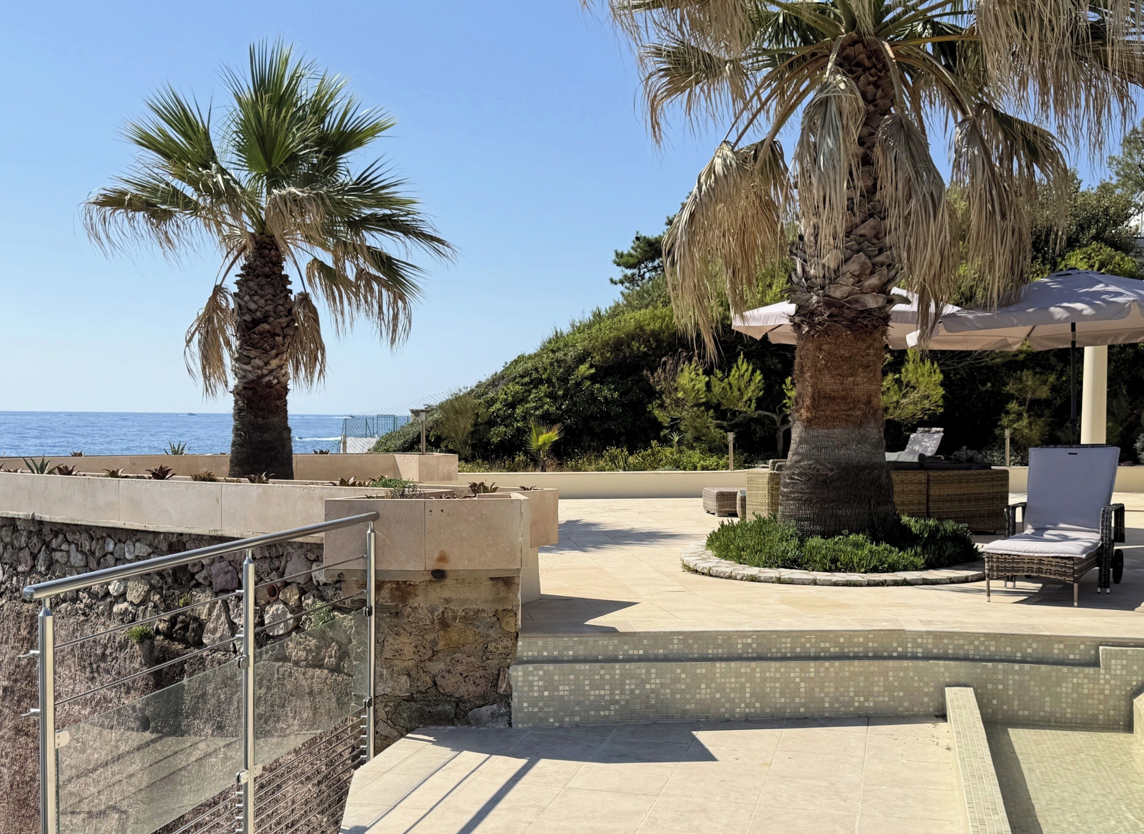 Un patio con vistas al mar, palmeras, mobiliario exterior y una pared de piedra.
