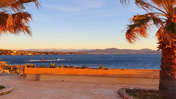 A coastal view with palm trees, a paved walkway, and a distant cityscape. - Superb villa "feet in the water"  (Roquebrune-Sur-Argens)