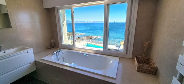Un baño con bañera, ventana con vista a una piscina y al mar, y suelo de baldosas.