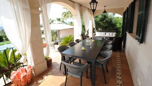 Un'area pranzo all'aperto riparata, con tavolo e sedie, un lampadario sospeso e vista su piante e piscina.