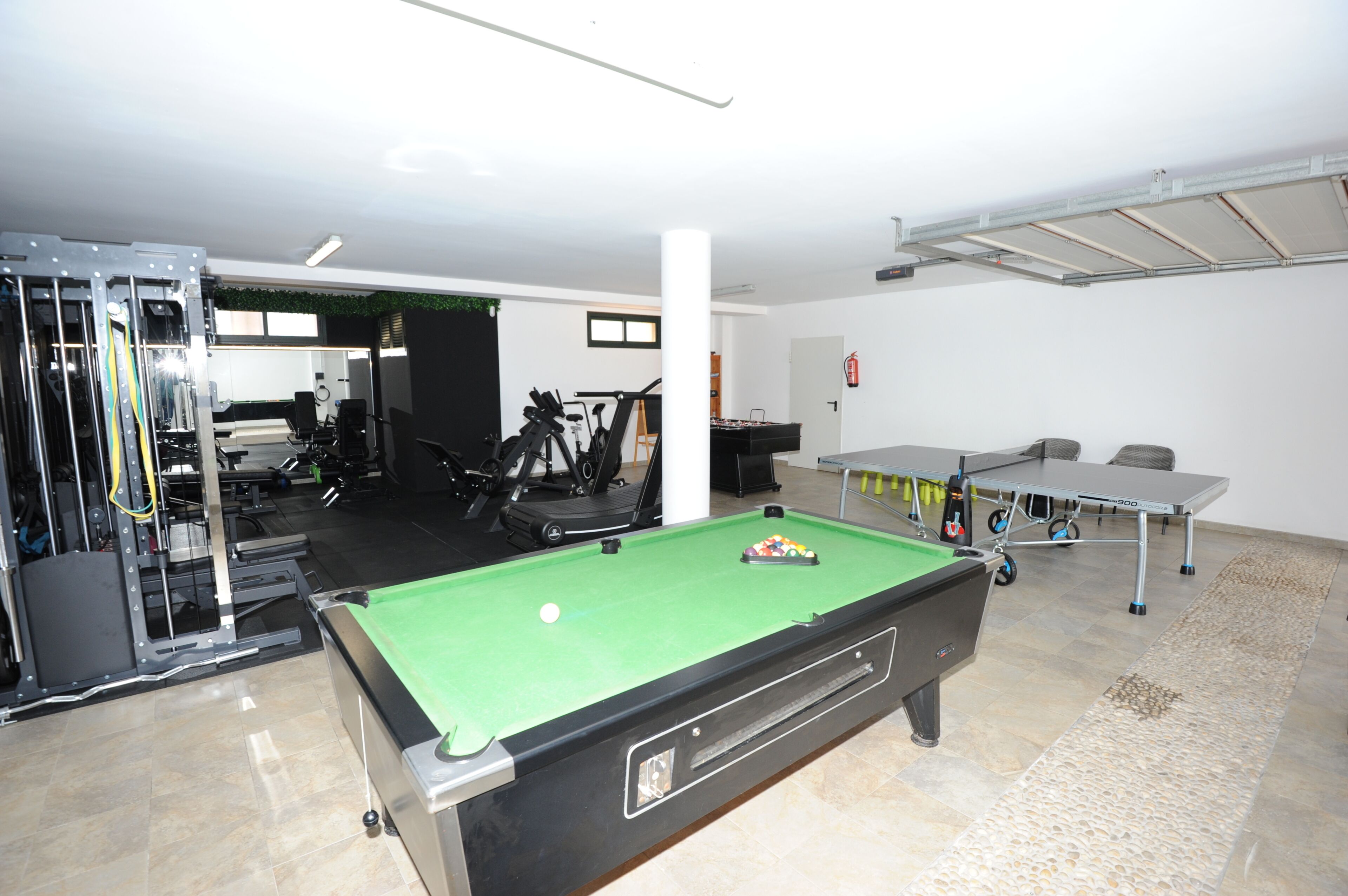 A pool table, exercise equipment, and a small table with chairs in a room with white walls and a tiled floor.