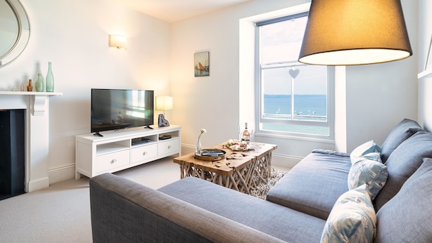 Ein modernes Wohnzimmer mit Fernseher, einem Holztisch, einer Couch und einem großen Fenster mit Meerblick.