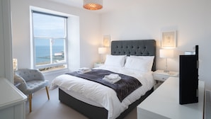 Ein modernes Schlafzimmer mit einem großen Bett, zwei Nachttischen, einem Sessel und Meerblick.
