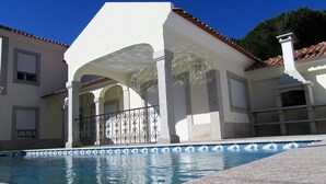A swimming pool with a tiled edge, a covered patio area with columns, and a building with a tiled roof. - Recent house with lagoon views 100 meters from the beach (LAGOA DE ALBUFEIRA)