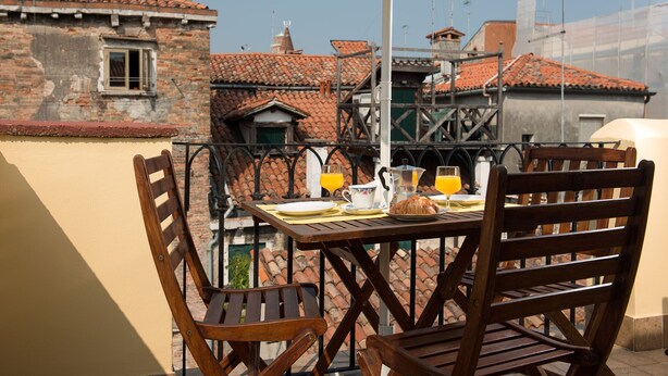 Un tavolo di legno con sedie, un ombrellone bianco e prodotti per la colazione su un balcone con vista su un paesaggio urbano caratterizzato da tetti in tegole rosse.