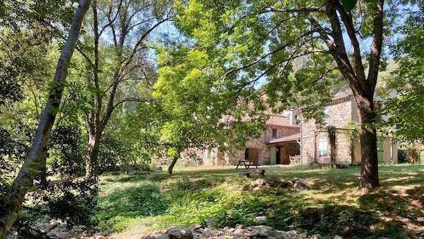 A rustic stone building with a stone roof, surrounded by a natural landscape with trees and a small stream.