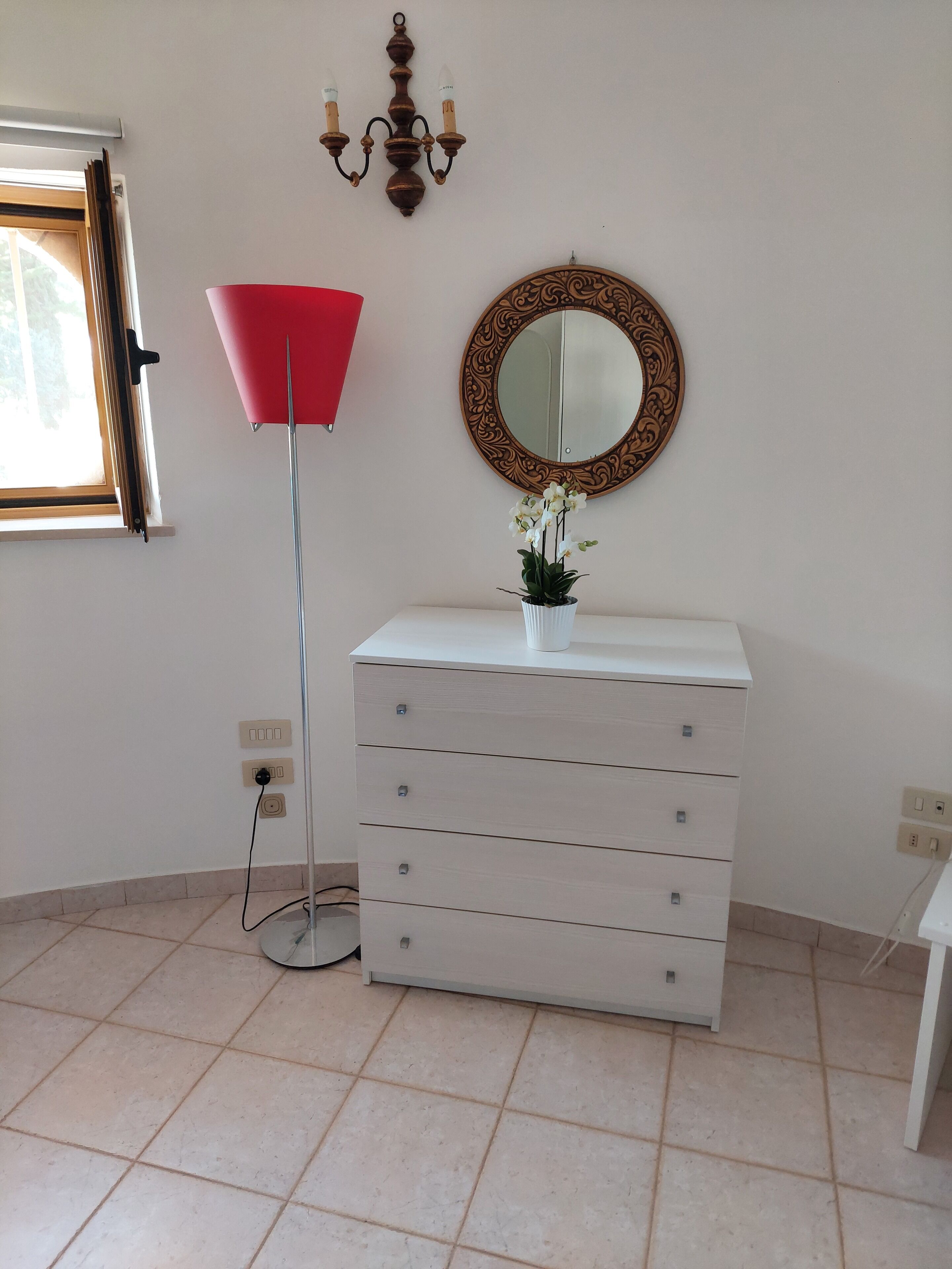 Una stanza con una cassettiera bianca, una lampada da terra rossa, uno specchio con cornice decorata e una pianta in vaso.