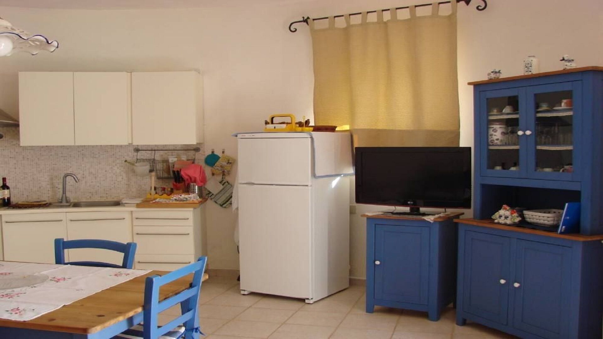 A compact kitchen with white cabinets, a white refrigerator, a blue cabinet with glass doors, a dining table with blue chairs, and a TV on a stand.