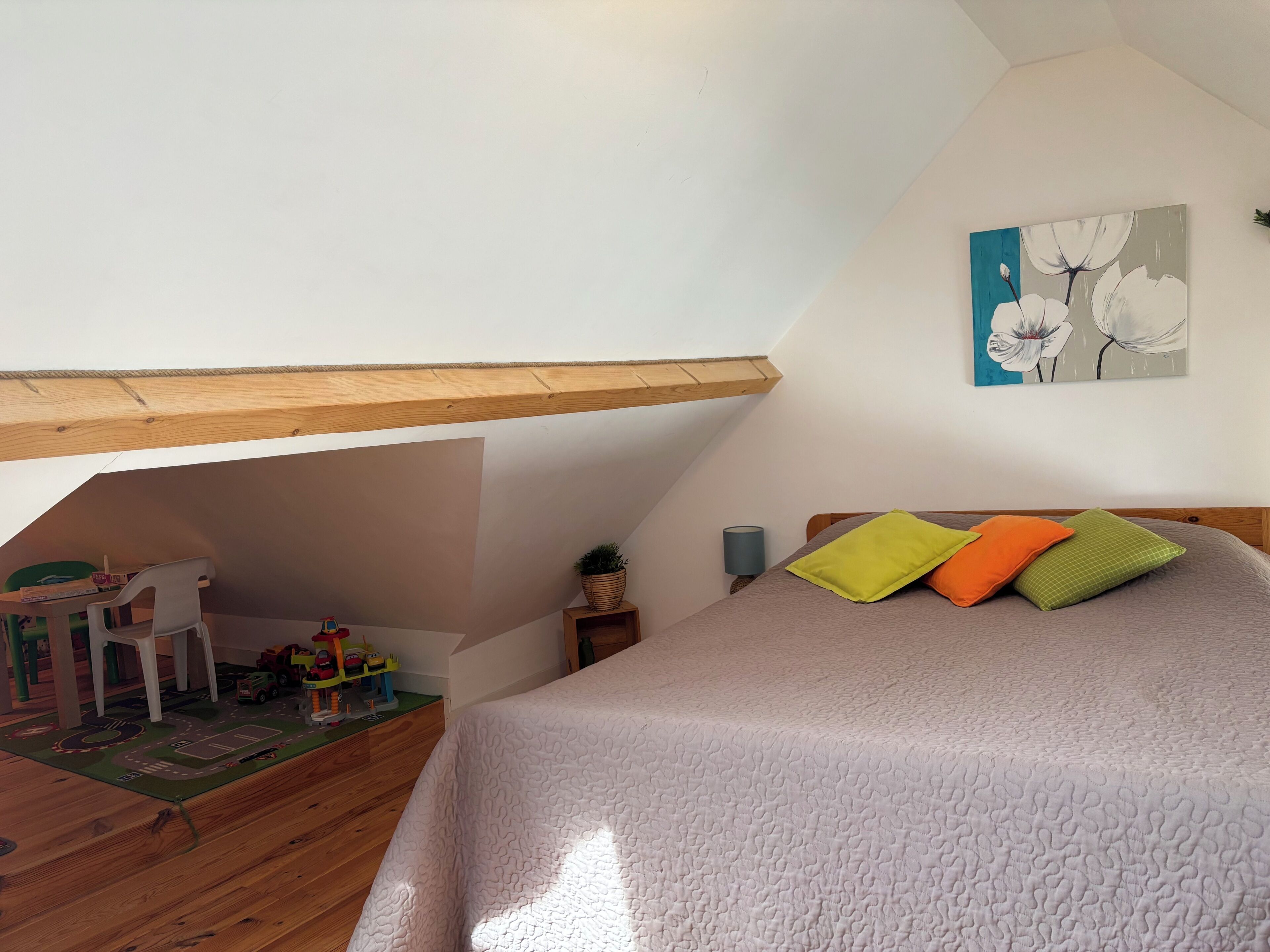 Une chambre avec un plafond en pente, un lit avec des oreillers, un petit bureau avec des jouets et un tableau accroché au mur.