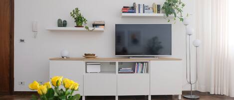 Un salon moderne avec un téléviseur à écran plat, des étagères blanches ornées de livres et de plantes, une table basse en bois et un vase rempli de fleurs jaunes.