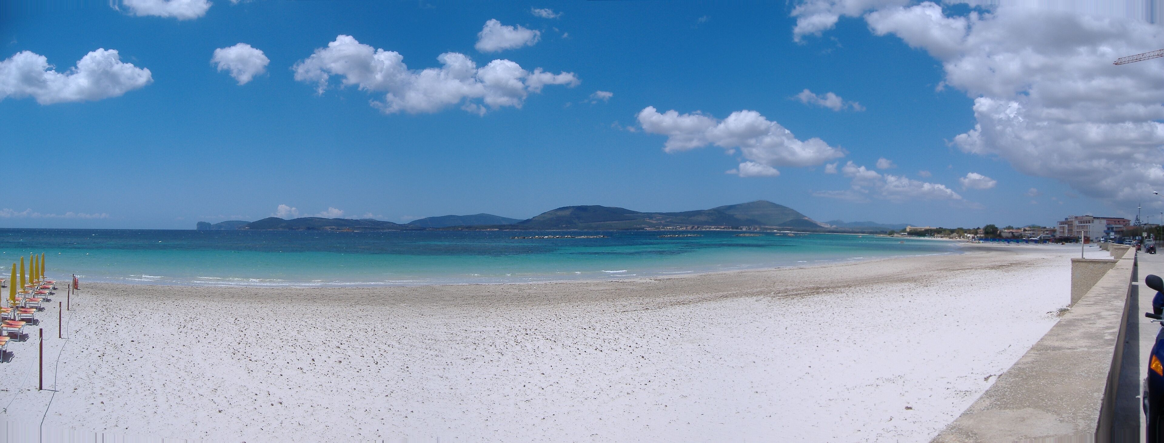 Een strand met wit zand, helderblauw water en een bergketen in de verte.