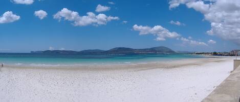 Een strand met wit zand, helderblauw water en een bergketen in de verte.