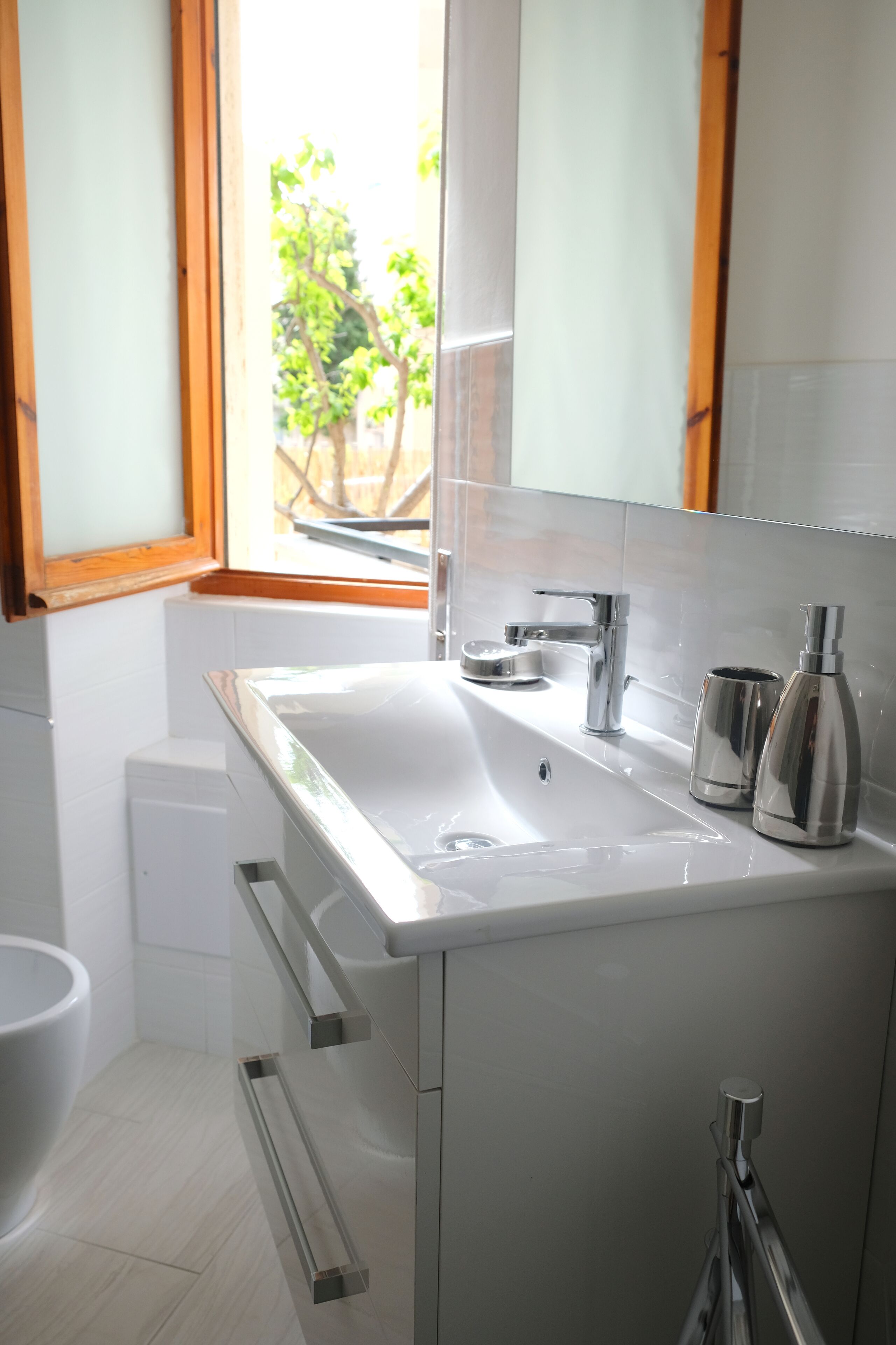 A modern bathroom with a white sink, chrome faucet, and a large mirror. There is a window above the sink, and a toilet is visible to the left.