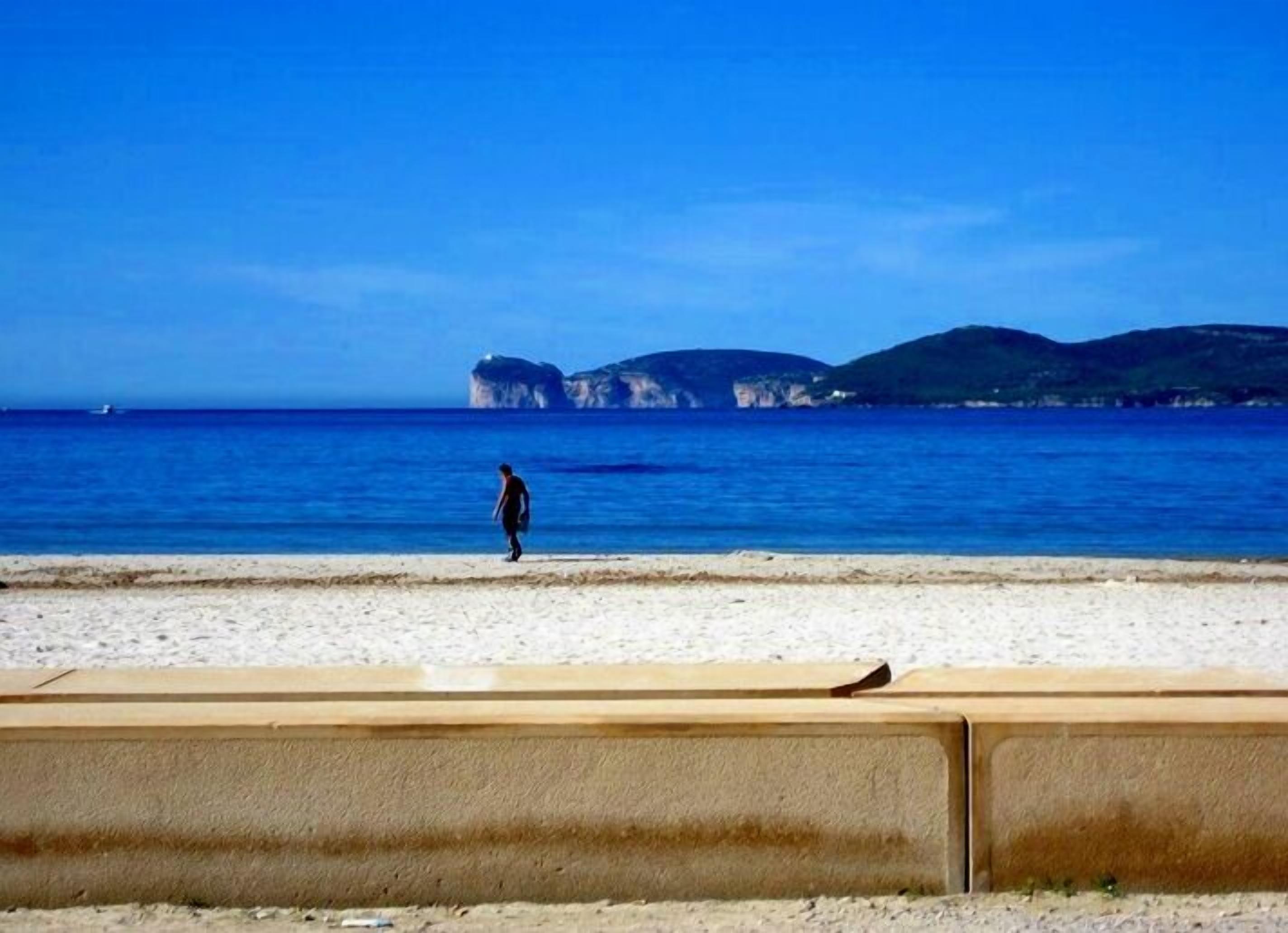 Una playa con una persona caminando, cielo despejado y acantilados a lo lejos.