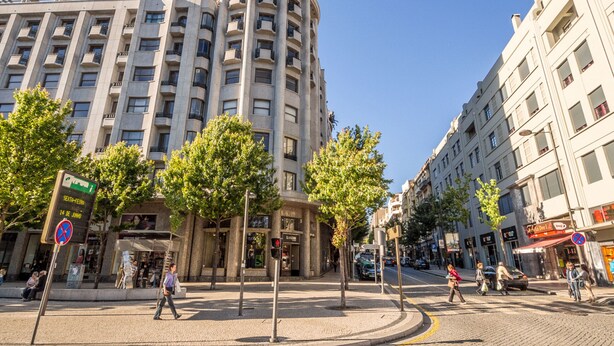 A multi-story building with a curved corner, a pedestrian crossing, and trees lining the street. - Feel Porto Downtown Heaven (Oporto)