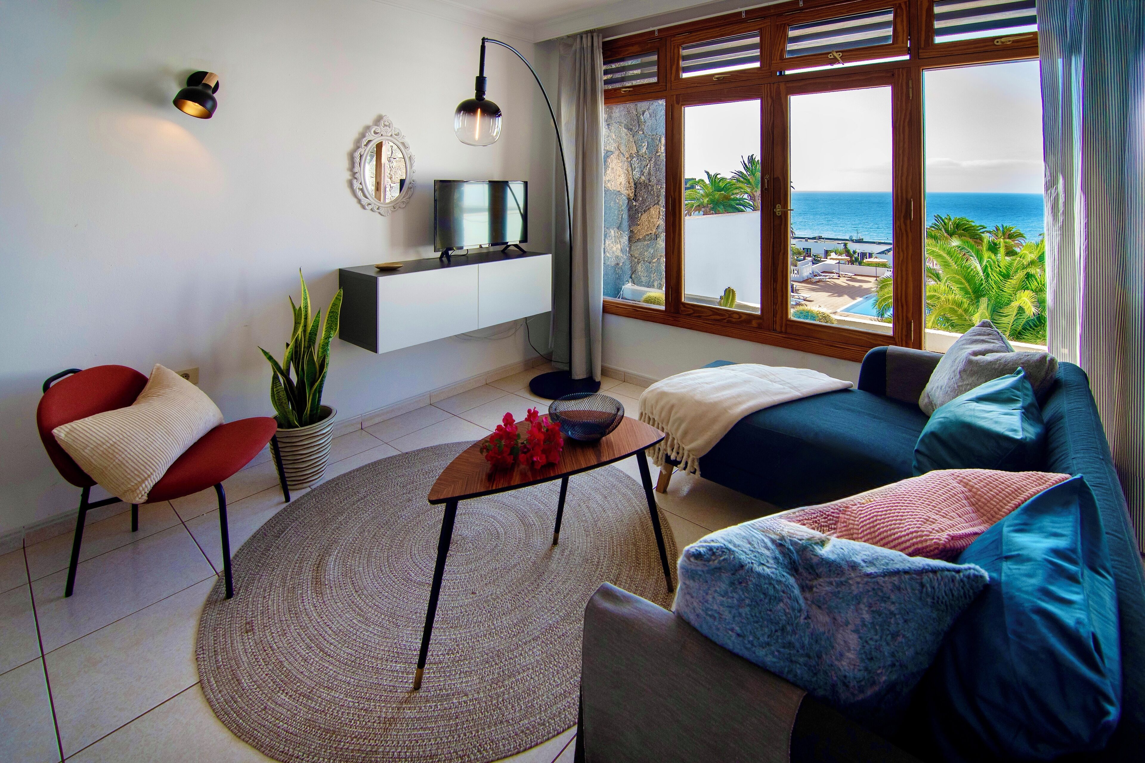 Un salon moderne avec un téléviseur posé sur une étagère blanche, un fauteuil rouge, un canapé bleu et une vue sur l’océan par de grandes fenêtres.