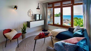 A modern living room with a TV on a white shelf, a red chair, a blue sofa, and a view of the ocean through large windows.
