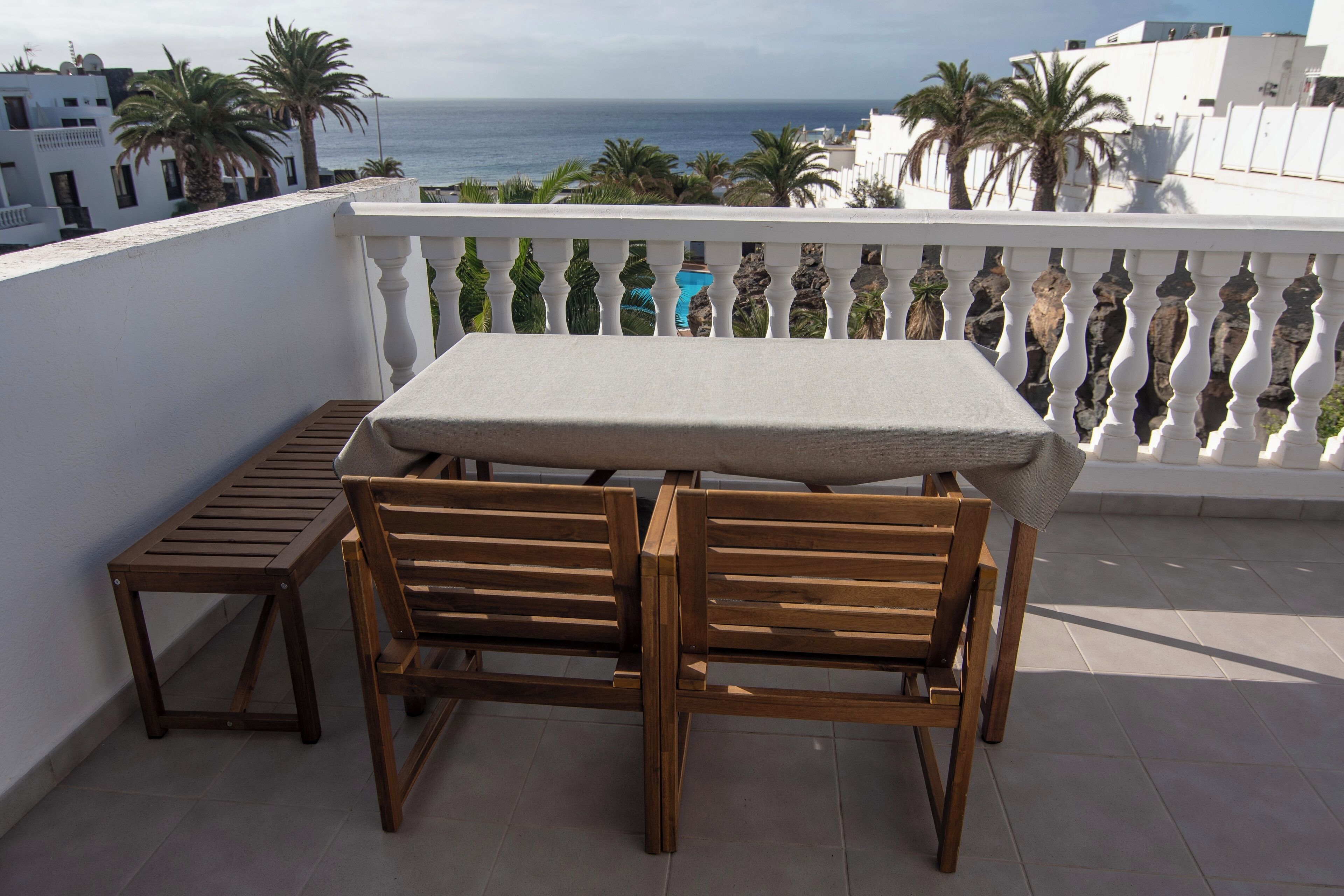 Un balcón con una mesa y dos sillas de madera, con vista al mar, palmeras y edificios.