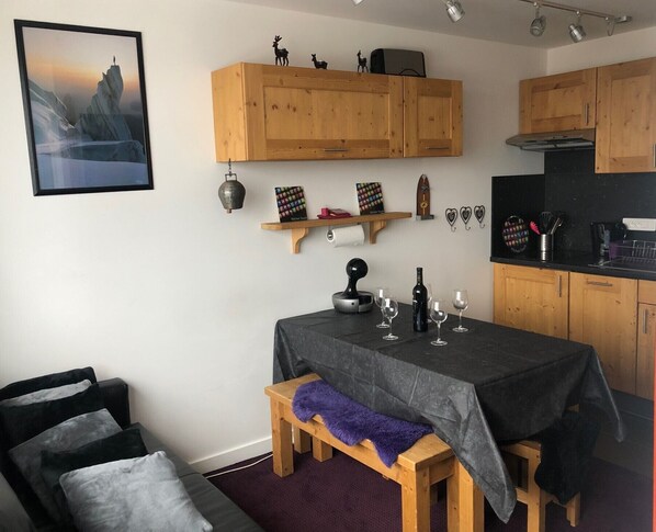 A cozy kitchen with wooden cabinets, a dining table with wine glasses, and a bench with a purple throw. - VERY NICE 2-ROOM APARTMENT FOR 4/5 PEOPLE, RECENTLY REFURBISHED (Morzine)