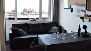 A modern living room with a black leather sofa, a dining table with wine glasses, and a view of the city through large windows. - VERY NICE 2-ROOM APARTMENT FOR 4/5 PEOPLE, RECENTLY REFURBISHED (Morzine)