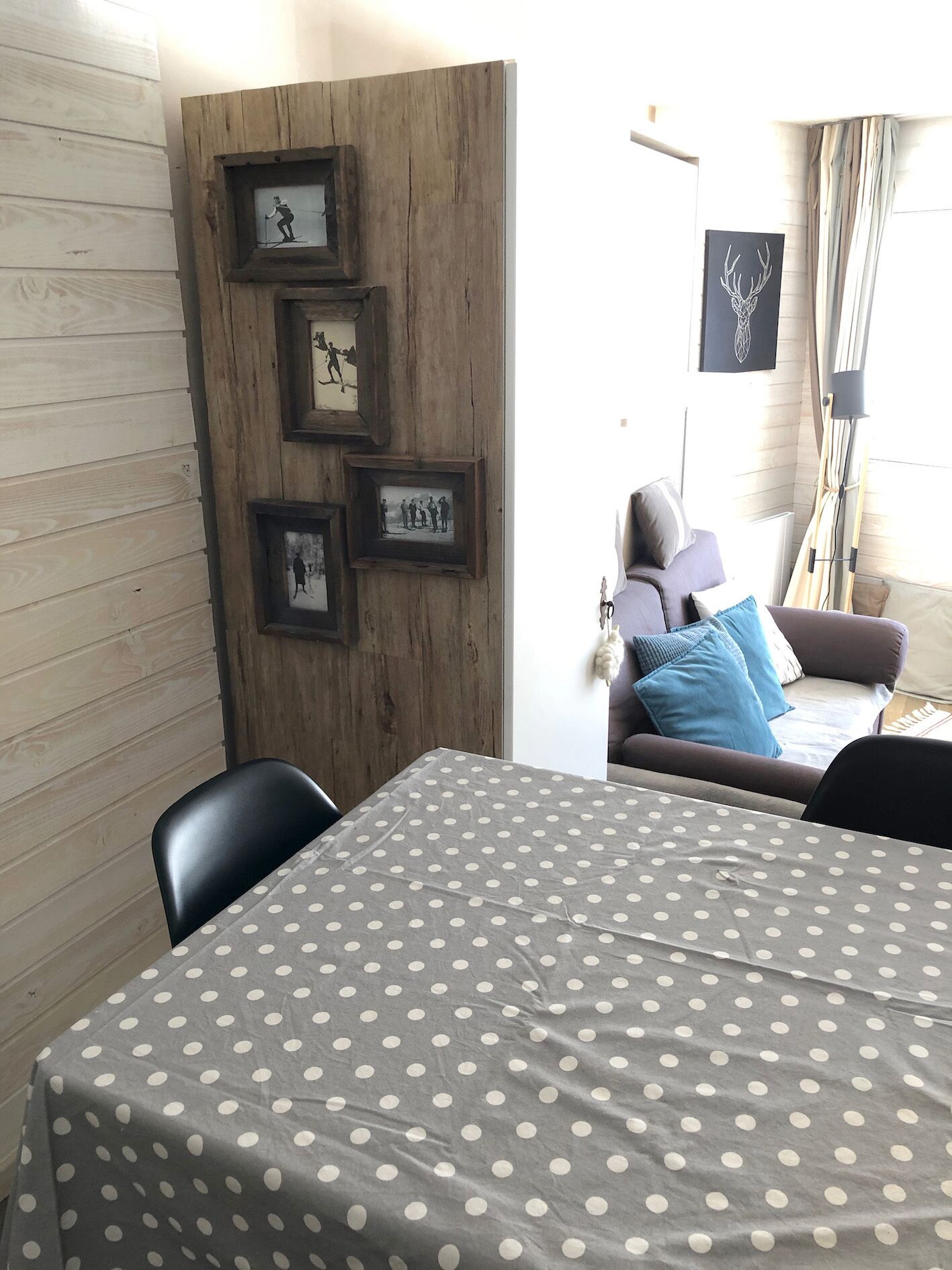 Una habitación con pared de madera, una mesa de comedor con mantel de lunares, un sofá con cojines azules y blancos, y cuadros en la pared.