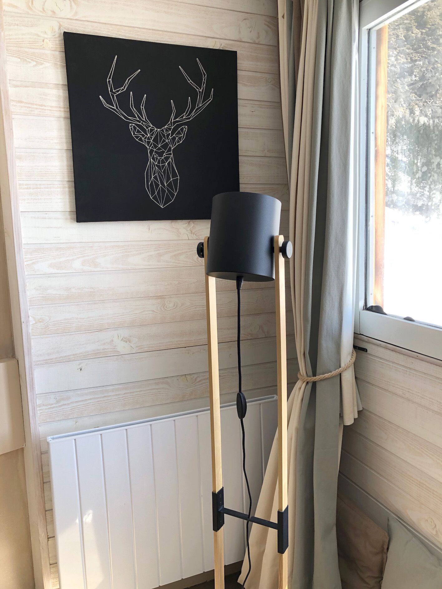 A room with a wooden wall, a black artwork of a deer head, a floor lamp, and a window with curtains.