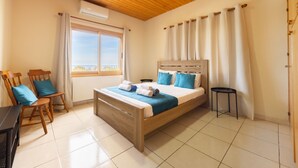 Een slaapkamer met een houten bed, een bureau met stoelen, een raam met gordijnen en uitzicht op de lucht.