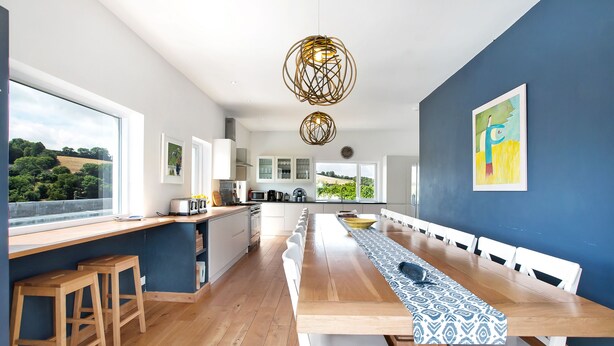A modern kitchen with a long wooden table, white chairs, and a view of the countryside through large windows. - Large Luxury House with Heated Pool, Hot Tub, Cinema close to coast near Shaldon (Stokeinteignhead)