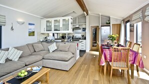 A modern living room with a grey sofa, a wooden coffee table with apples, and a dining area with a table set for two. - Luxury 3 bed Lodge In Soar, Salcombe, Devon, Sea & Country views (Salcombe)
