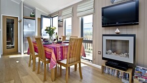 A dining area with a table set for four, a television mounted above a fireplace, and a bookshelf with DVDs. - Luxury 3 bed Lodge In Soar, Salcombe, Devon, Sea & Country views (Salcombe)