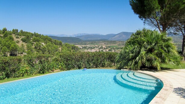 A clear blue swimming pool with a curved edge, surrounded by lush greenery and a view of distant hills and mountains.