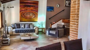 Ampla sala de estar com sofá, mesa de centro e mesa de jantar com cadeiras. Um grande quadro abstrato está na parede, e há uma escada que leva a outro andar.