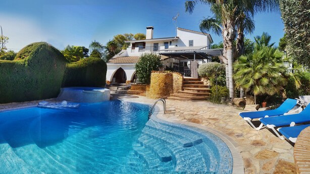 Piscina com deck de pedra, rodeada por espreguiçadeiras e um edifício com fachada branca e degraus de pedra.