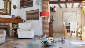 Uma sala de estar com lareira, uma poltrona branca, um abajur vermelho e um espaço de jantar com cadeiras de madeira.