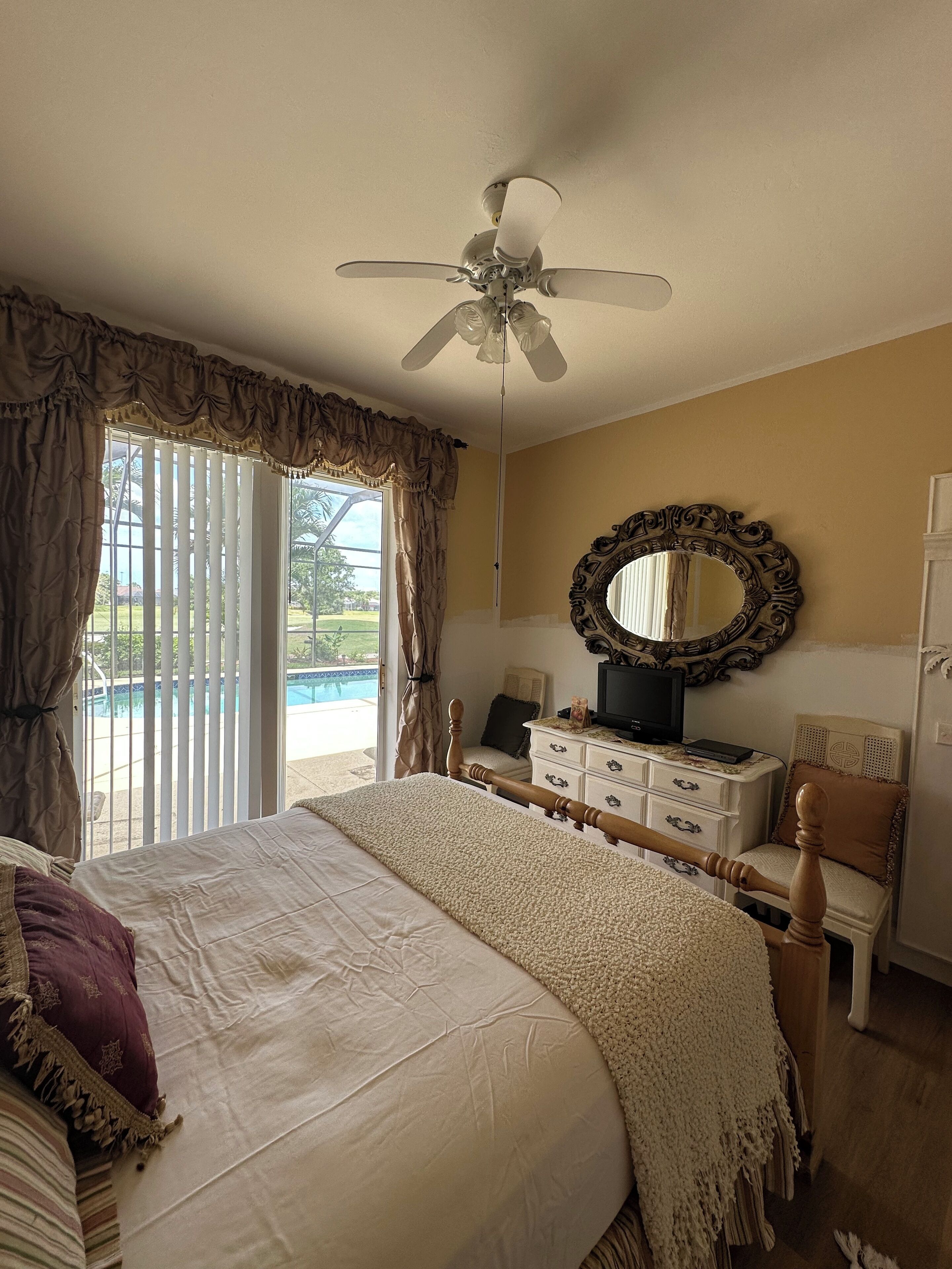 A bedroom with a large bed, a ceiling fan, a dresser, a mirror, and a view of a swimming pool.