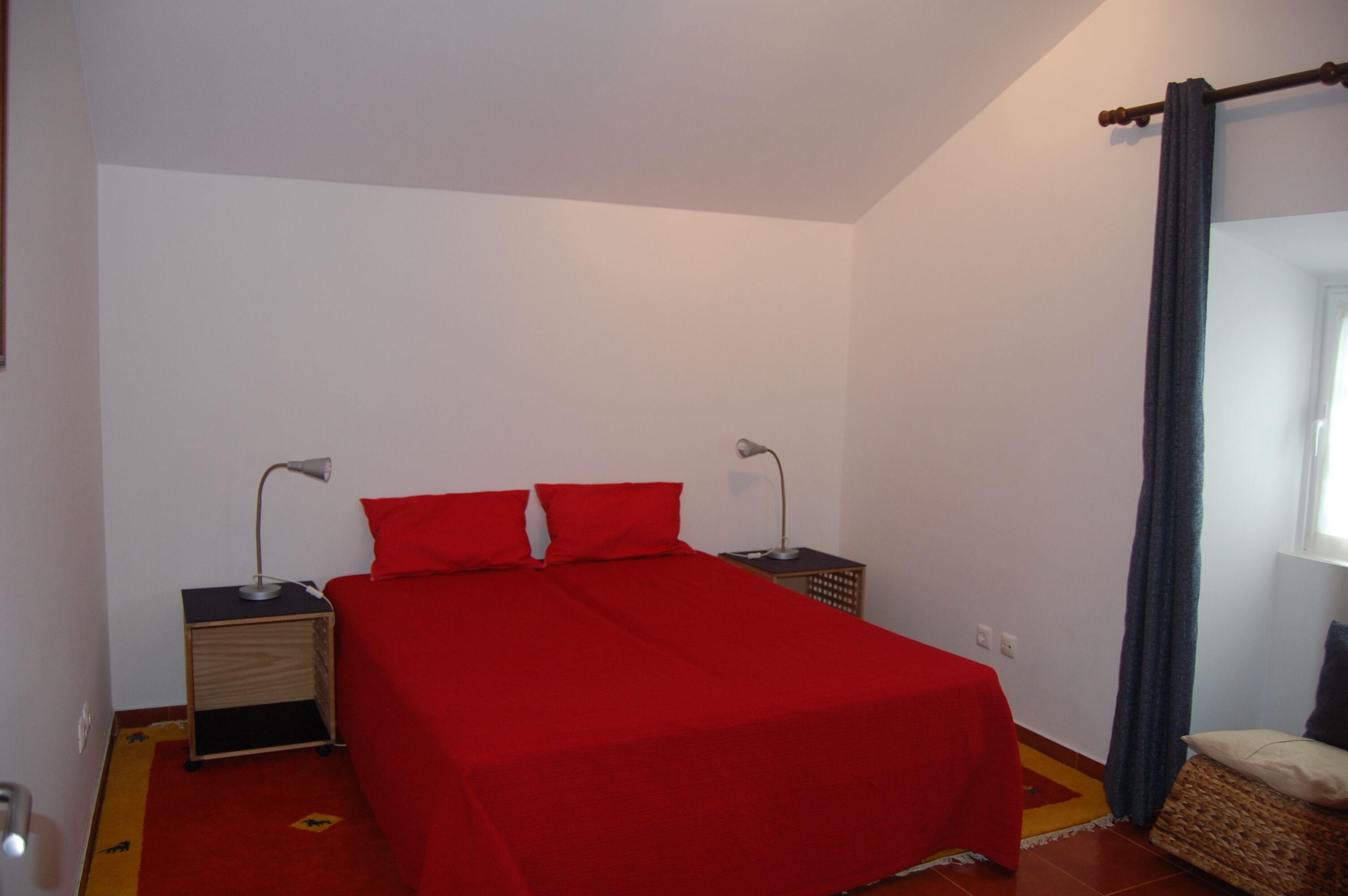 Un dormitorio con una colcha roja, dos lámparas de noche, una mesita auxiliar pequeña, una silla de mimbre y una ventana con cortina.