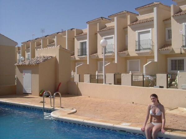 A swimming pool with a diving board, a woman sitting by the pool, and a building with balconies and windows in the background.