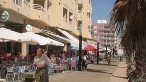 Eine Küstenpromenade mit Außenbestuhlung, einer Palme und einem mehrstöckigen Gebäude mit Balkonen.