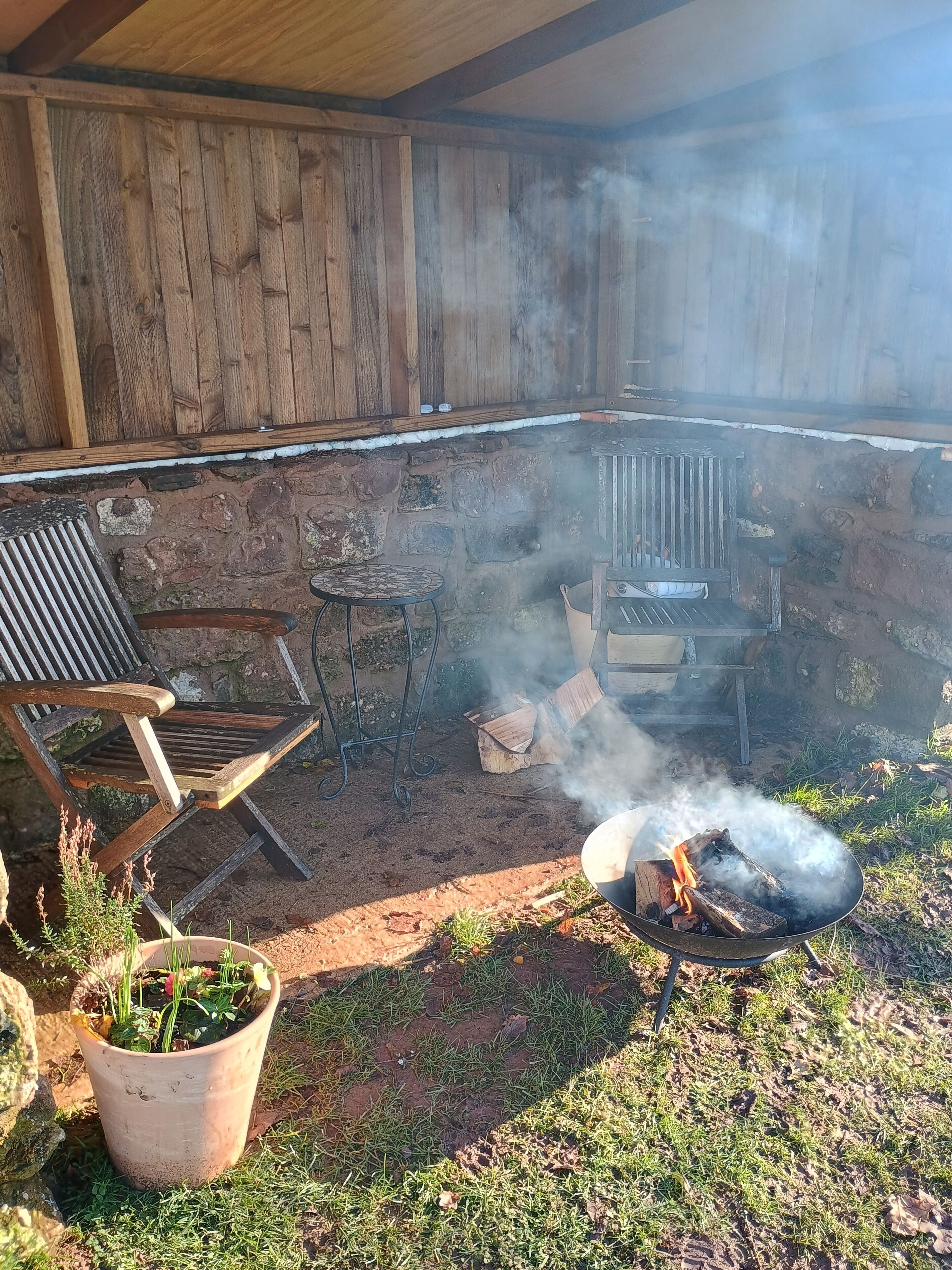 An outdoor patio with a wooden bench, a metal grill, and a fire burning.
