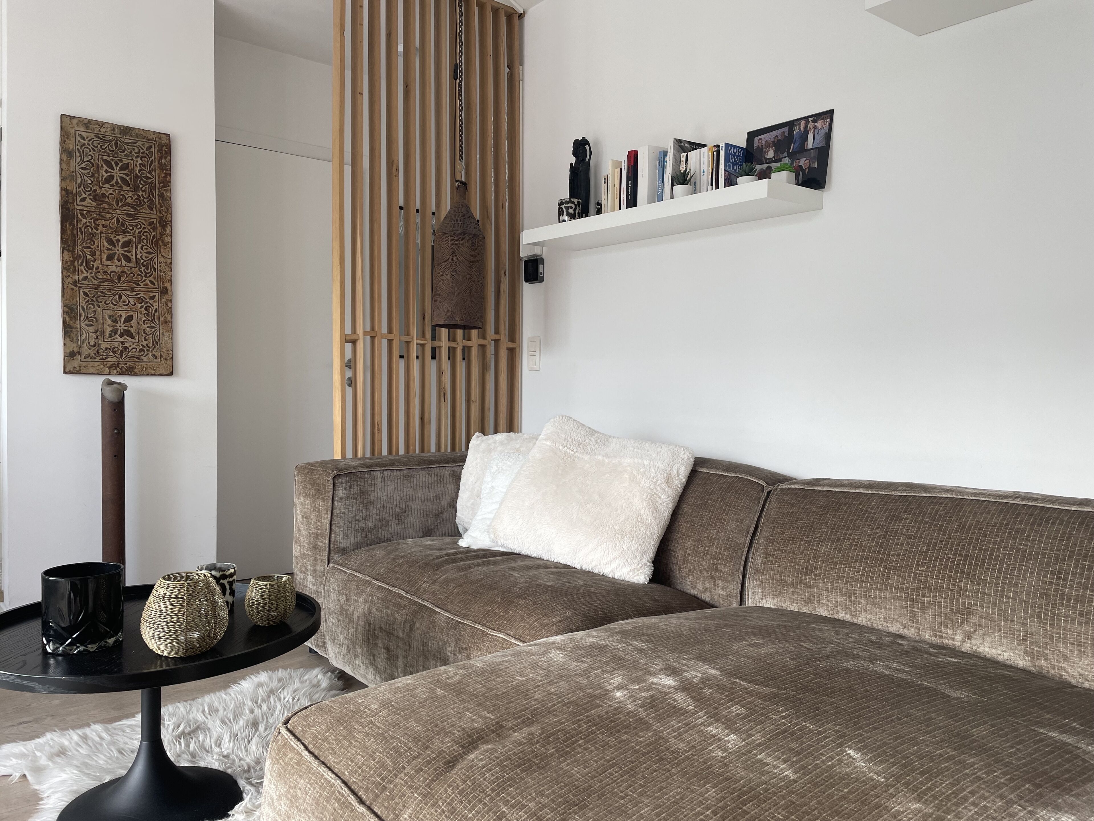 A modern living room with a grey sofa, a black coffee table with decorative items, a white furry rug, a wooden slatted partition, and a white shelf with books and a picture frame.