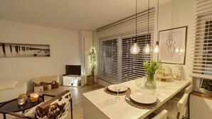 Ein modernes Wohnzimmer mit Essbereich, ausgestattet mit einem weißen Tisch für zwei Personen, einer Couch und einem Fernseher.