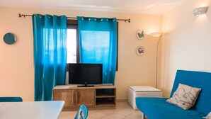 Une pièce de taille modeste, avec un téléviseur posé sur un meuble en bois, un canapé bleu agrémenté d’un coussin décoratif, une petite table blanche et une chaise bleue.