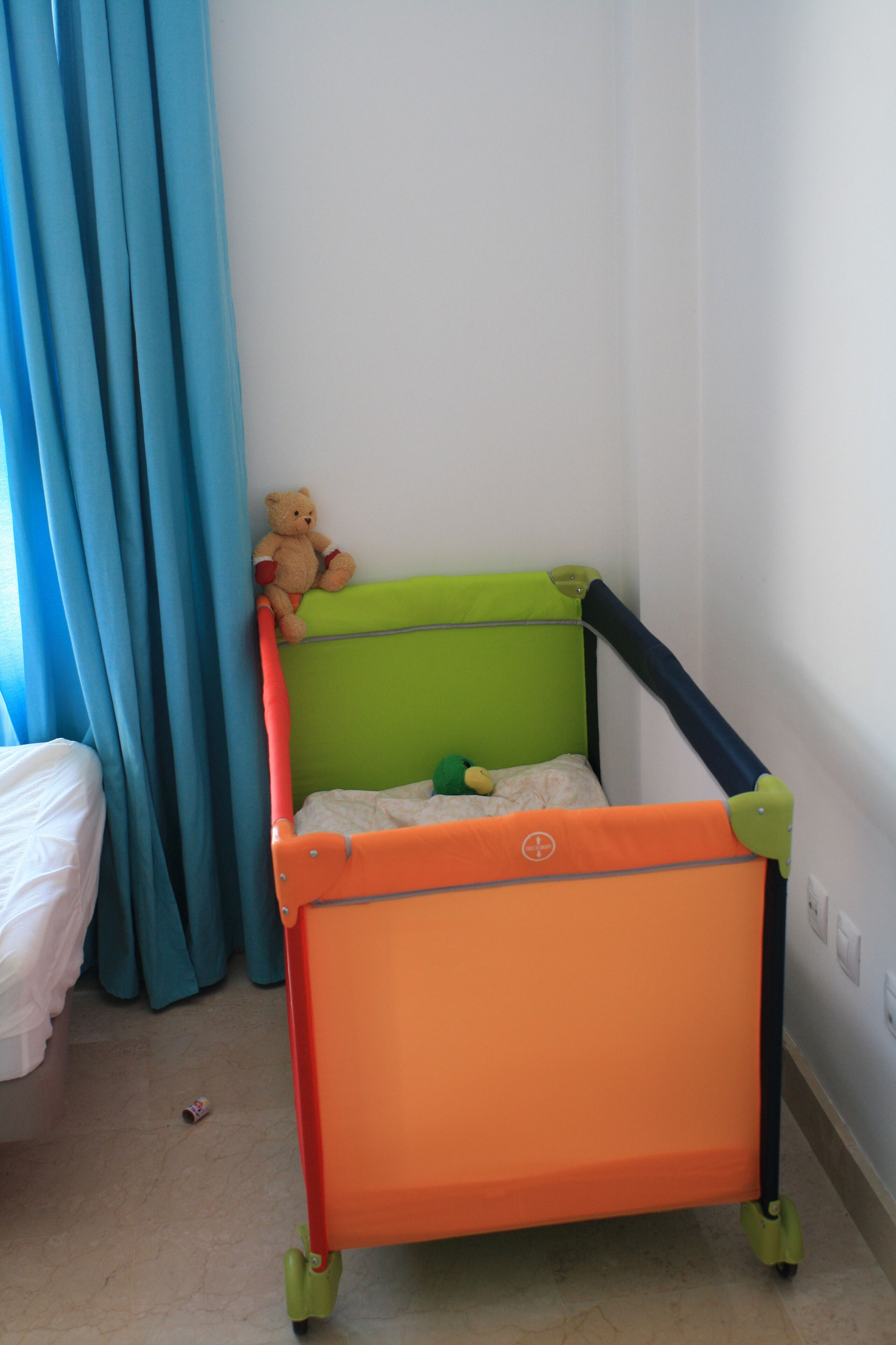 A child's playpen with a green top and orange bottom, a teddy bear on top, and a bed with white sheets in the background.