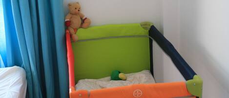 A child's playpen with a green top and orange bottom, a teddy bear on top, and a bed with white sheets in the background.