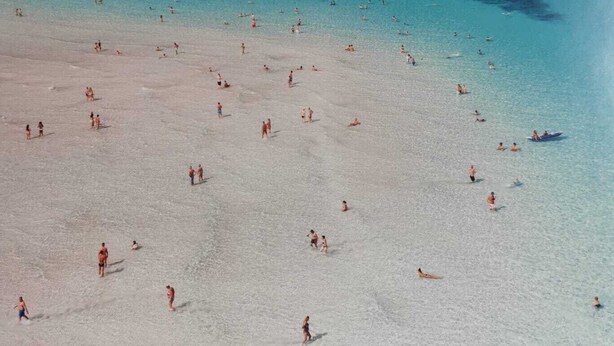 A beach with clear turquoise water and a sandy shore. There is a tower-like structure on a small island in the distance.
