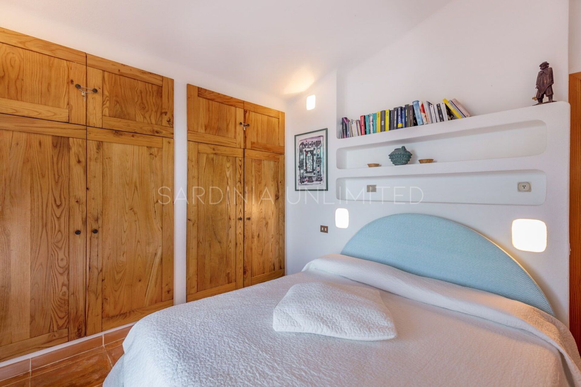 A bedroom with a bed, wooden wardrobe, and a shelf with books and decorative items.