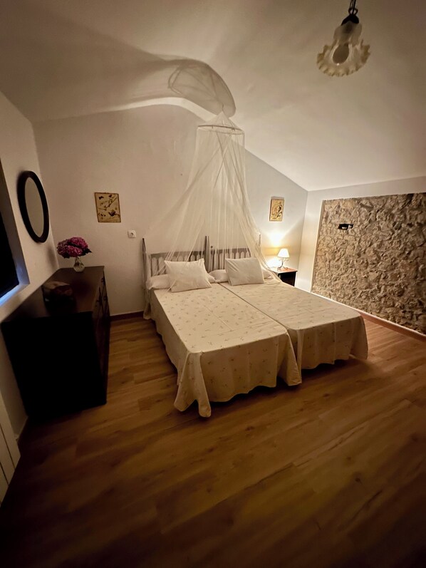 A bedroom with a double bed, a nightstand, a bedside table, a wall-mounted mirror, and a decorative vase with flowers.