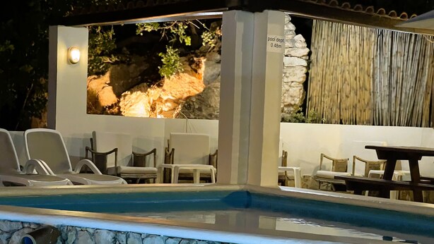 A pool area with lounge chairs and a table, surrounded by stone walls and a decorative rock feature.